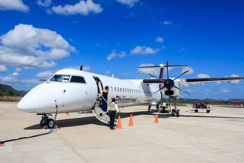 A small propeller airplane, ready to whisk passengers to El Nido, is parked on a runway under a clear blue sky. As travelers board via a stairway, two workers handle luggage nearby. The airplane proudly displays "Philippines" on the side, with orange cones placed around it for safety.