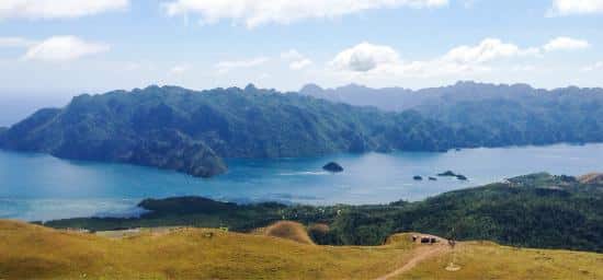 Scenic view of rolling green hills overlooking a serene blue bay, reminiscent of Coron, surrounded by rugged, forested mountains under a partly cloudy sky. Small islands dot the calm water, creating a picturesque and tranquil landscape perfect for an unforgettable boat tour.