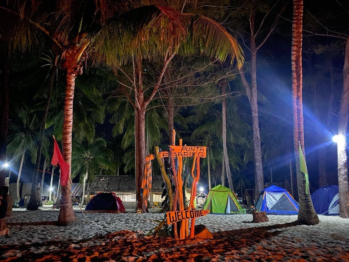 Under the starlit sky, the night beach camping scene at Punta Baring unfolds with tents nestled among palm trees. A wooden sign warmly reads "Welcome," while nearby, whispers of island hopping and tales from a Balabac boat tour invite adventurers to explore further.