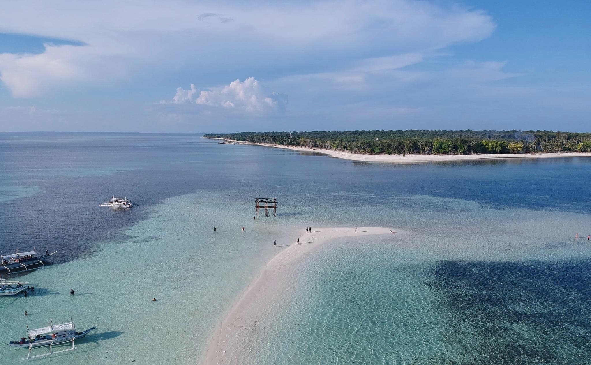 Aerial view of a sandy beach in Balabac with clear blue water, scattered boats perfect for island hopping, and a wooden platform. Lush greenery lines the shore under a clear sky.