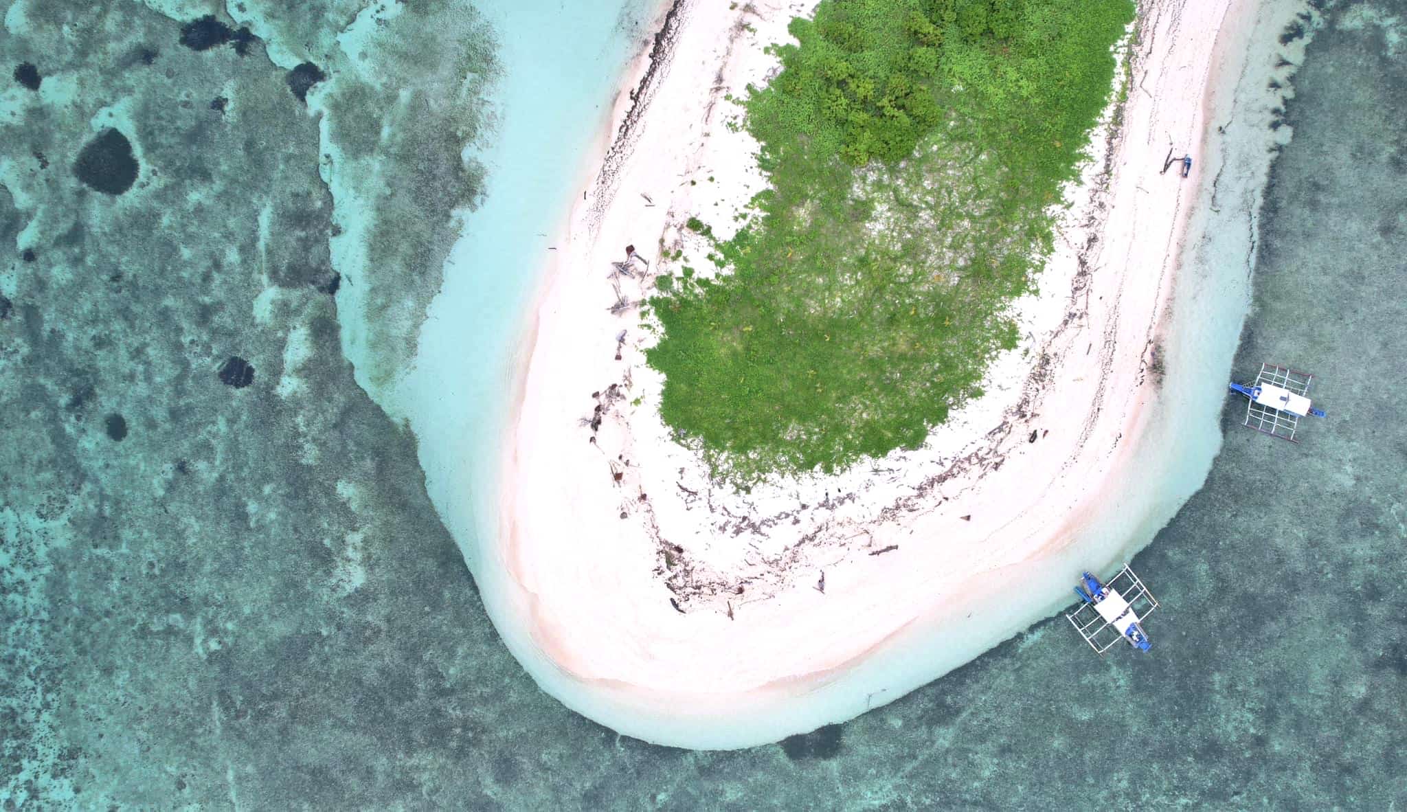 Aerial view of a small sandy island with lush vegetation, nestled in the clear turquoise waters of Balabac. Nearby, two boats hint at exciting island hopping adventures waiting to unfold.