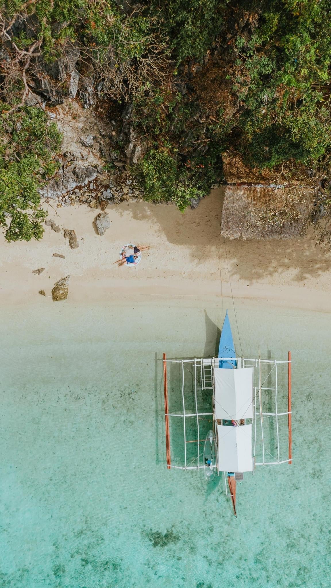 Aerial view of a white boat from a nearby island hopping adventure docked on Banol Beach’s sandy shore with turquoise waters. Two people sit near the shoreline beneath a tree, next to a small wooden hut. Dense green foliage envelopes the area.