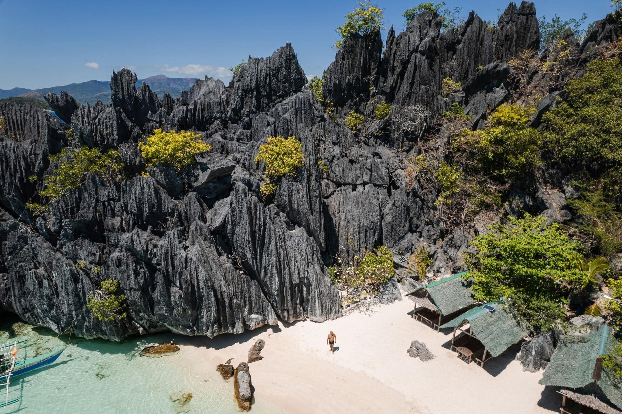 A person stands on Banol Beach's white sandy shore, surrounded by dramatic, jagged black rock formations. Two small huts with green roofs sit nearby. Clear turquoise water laps at the shoreline as lush greenery is interspersed among the rocks, making it an ideal spot for island hopping.