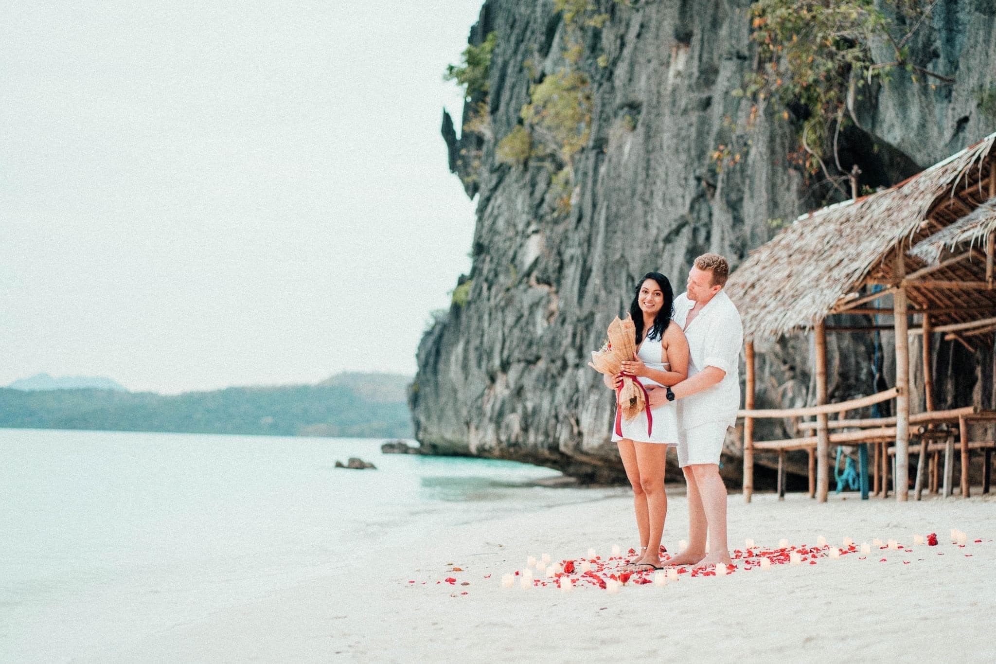 A couple stands on the sandy shores of Banol Beach near a bamboo hut and rocky cliffside. The man holds the woman, who is clutching a bouquet, while red petals are scattered around them. An ocean awaits in the background, perfect for island hopping adventures.