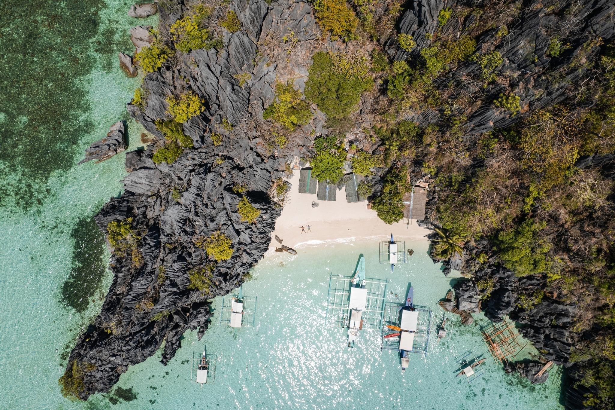 Aerial view of Banol Beach with clear turquoise water. Several small boats, perfect for island hopping, are anchored near the shore. The secluded beach is surrounded by rocky cliffs and lush greenery, with a few people visible on the sand.