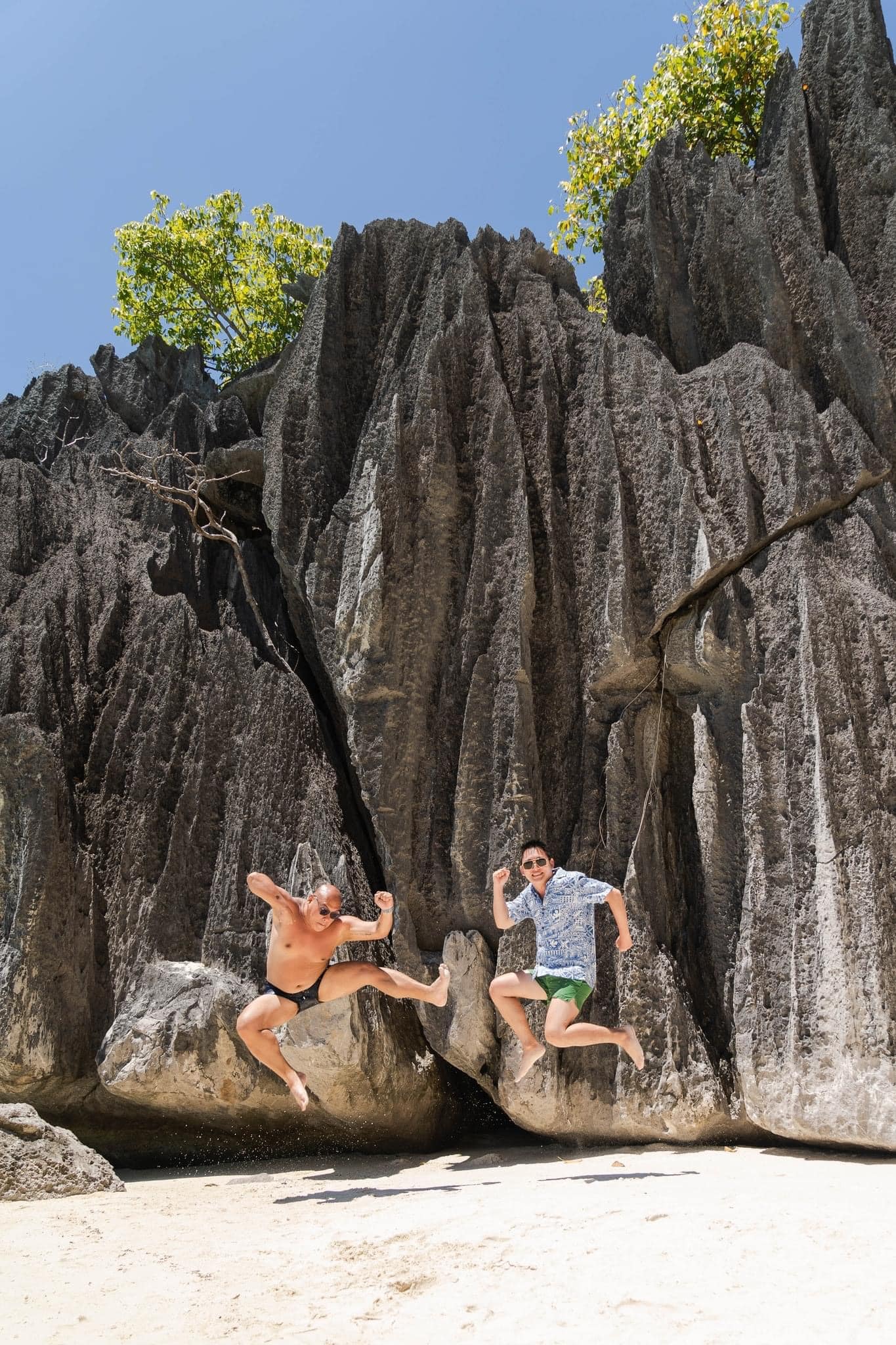 Two people are jumping playfully in the air on a sandy Banol Beach, framed by towering jagged rock formations. One is shirtless, and the other wears a shirt and shorts. A clear blue sky stretches overhead, inviting thoughts of island hopping adventures just beyond the horizon.