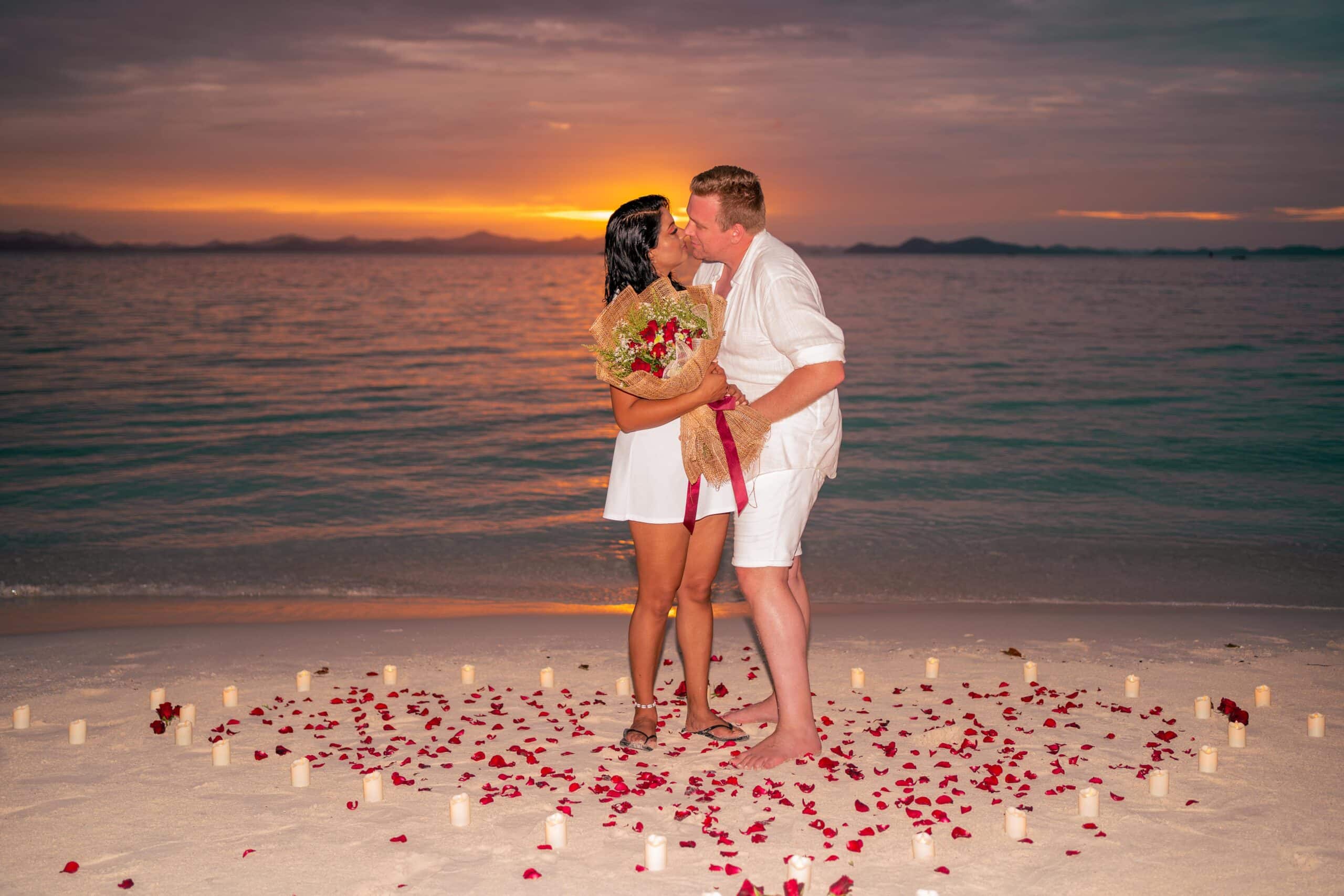 A couple kisses on a beach at sunset, standing on sand surrounded by scattered red rose petals and candles after a day of island hopping. The woman holds a bouquet of flowers as the ocean and an orange-hued sky over El Nido provide a romantic backdrop.