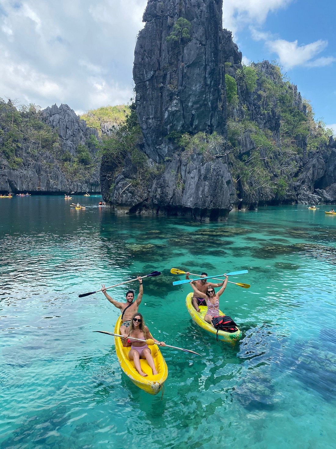 Three people in swimsuits paddle yellow kayaks in the clear turquoise waters of Big Lagoon, surrounded by dramatic limestone cliffs and lush greenery under a partly cloudy sky. They smile and raise their arms, fully immersed in the scenic island-hopping adventure near El Nido.