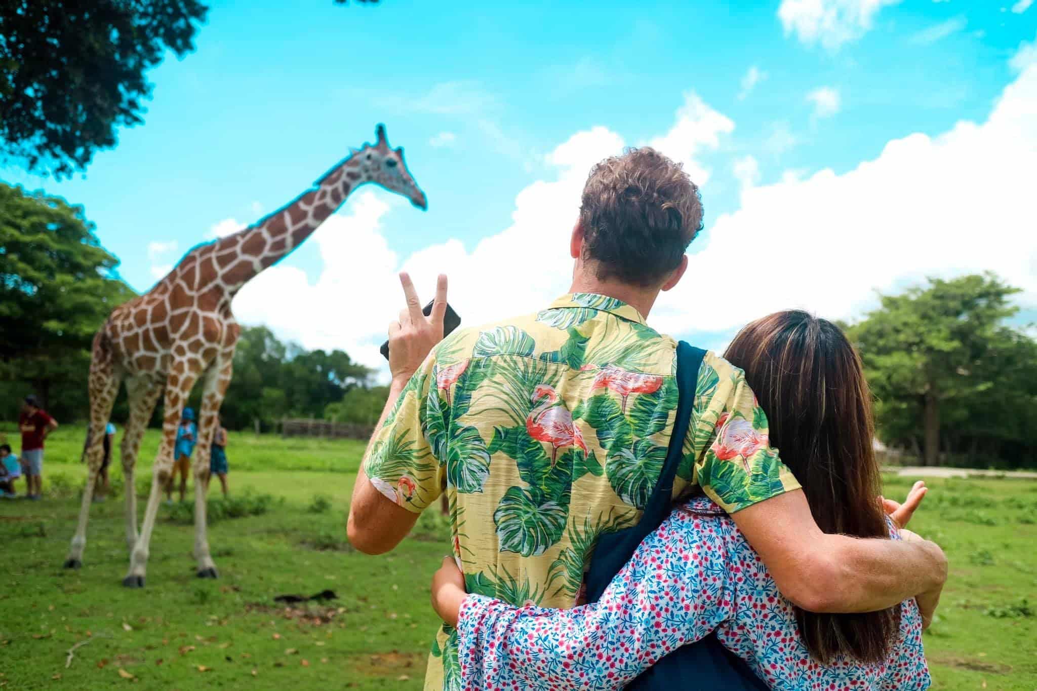 A couple stands with their backs to the camera, gazing at a giraffe in the lush, green setting of Calauit Safari Park. The man wears a colorful shirt, flashing a peace sign, while the woman embraces him. Above them, the Palawan sky is bright with scattered clouds.