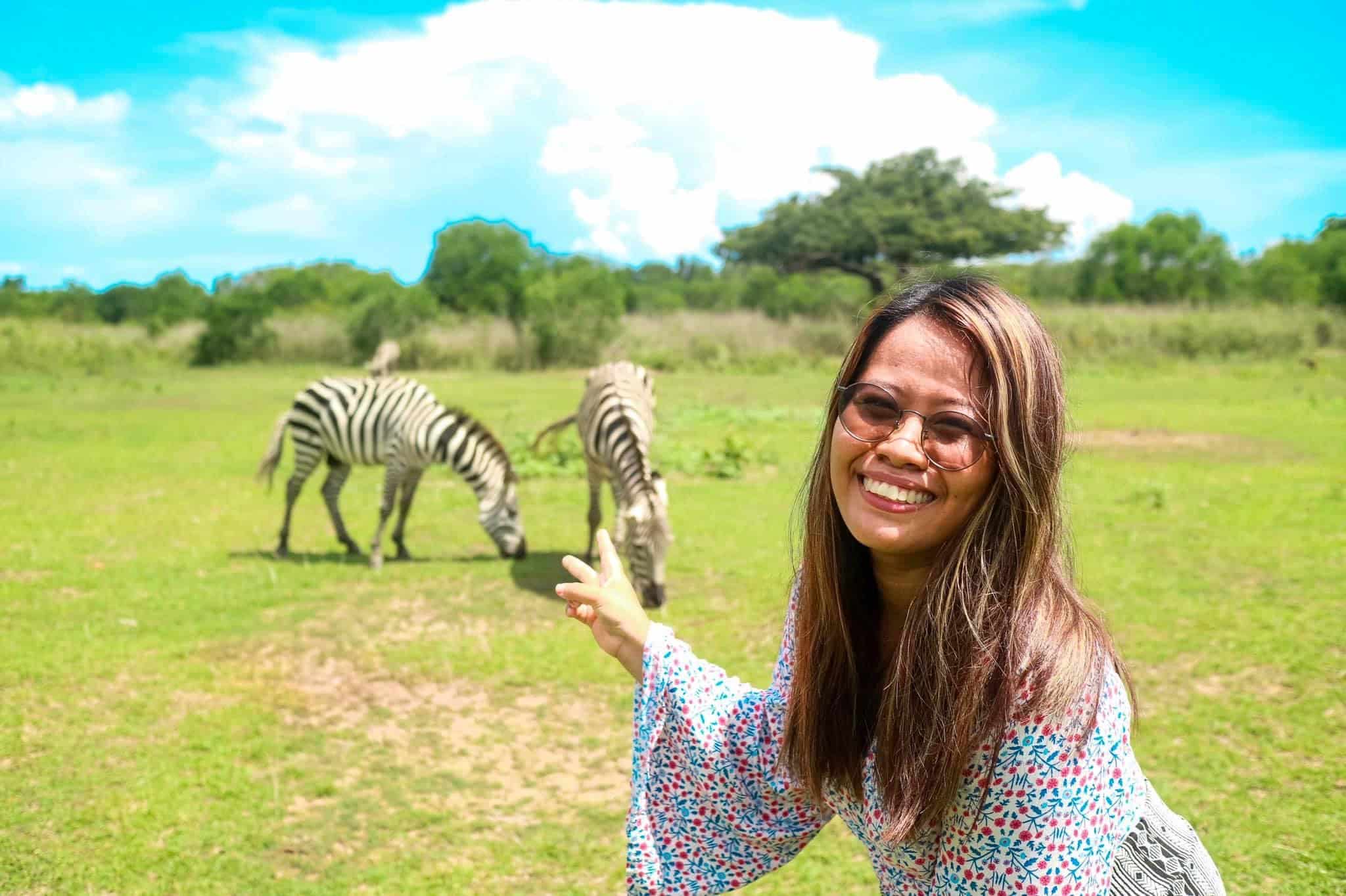A woman with long hair and glasses smiles and points towards two zebras grazing on a grassy field at Calauit Safari Park. The sky is bright blue with scattered clouds, and lush trees create a picturesque backdrop, reminiscent of the stunning landscapes of Palawan.