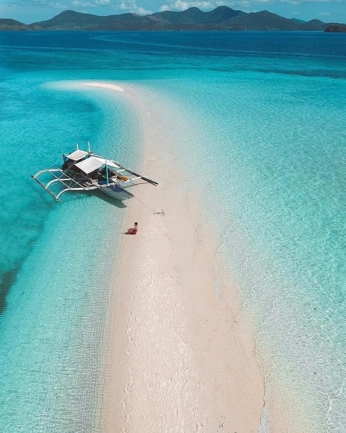 Aerial view of a small boat anchored on a narrow sandbar near Ditaytayan Island, surrounded by turquoise waters. A person is lying on the sandbar under the clear blue sky, with distant green hills of Coron, Palawan visible on the horizon.