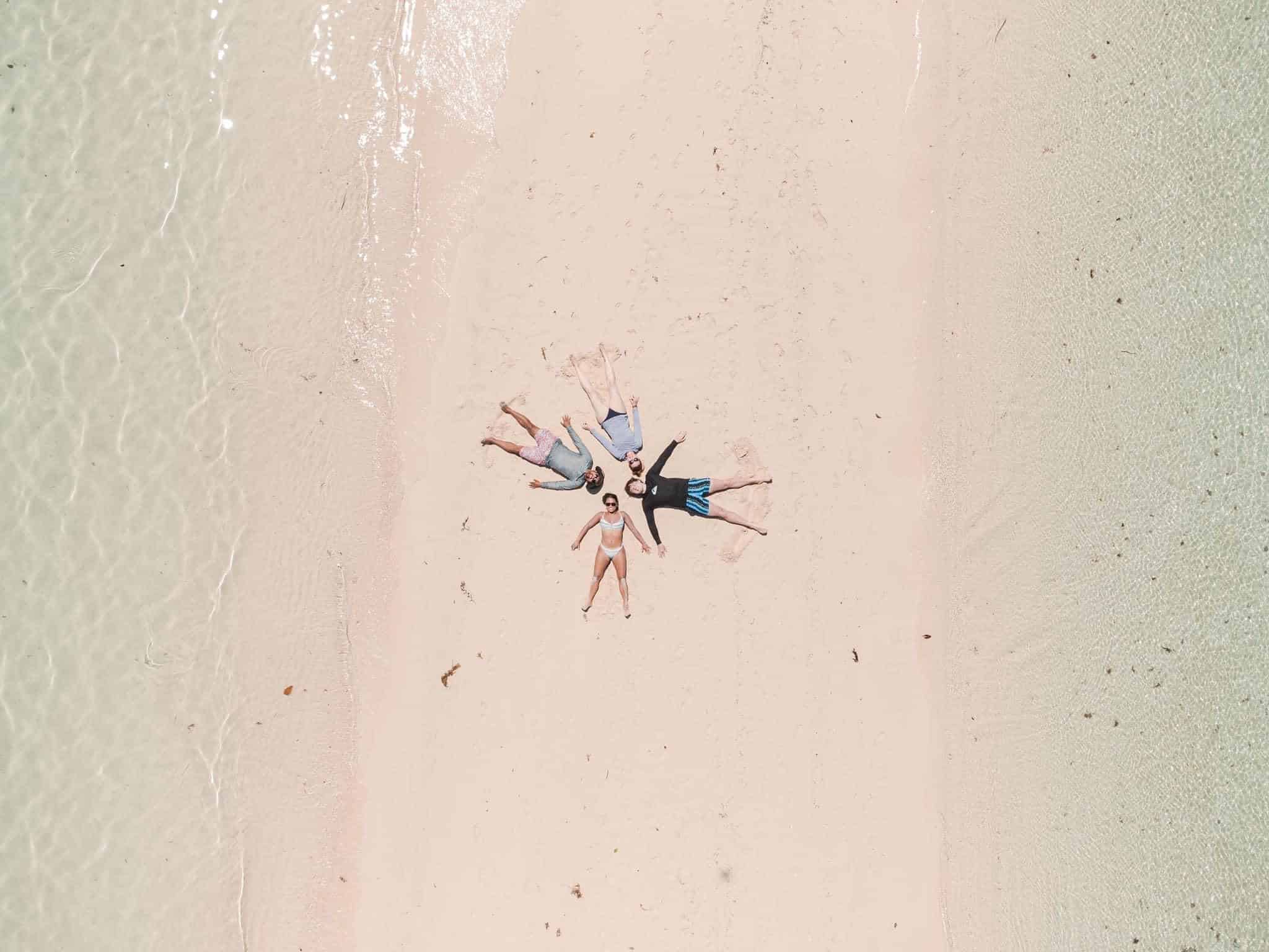 On Ditaytayan Island, a group of people lie in a circle on the sandy beach near Coron's clear, shallow water. Their heads are together, forming a star shape with their bodies. The photo, taken from above, captures this stunning scene in Palawan's picturesque paradise.