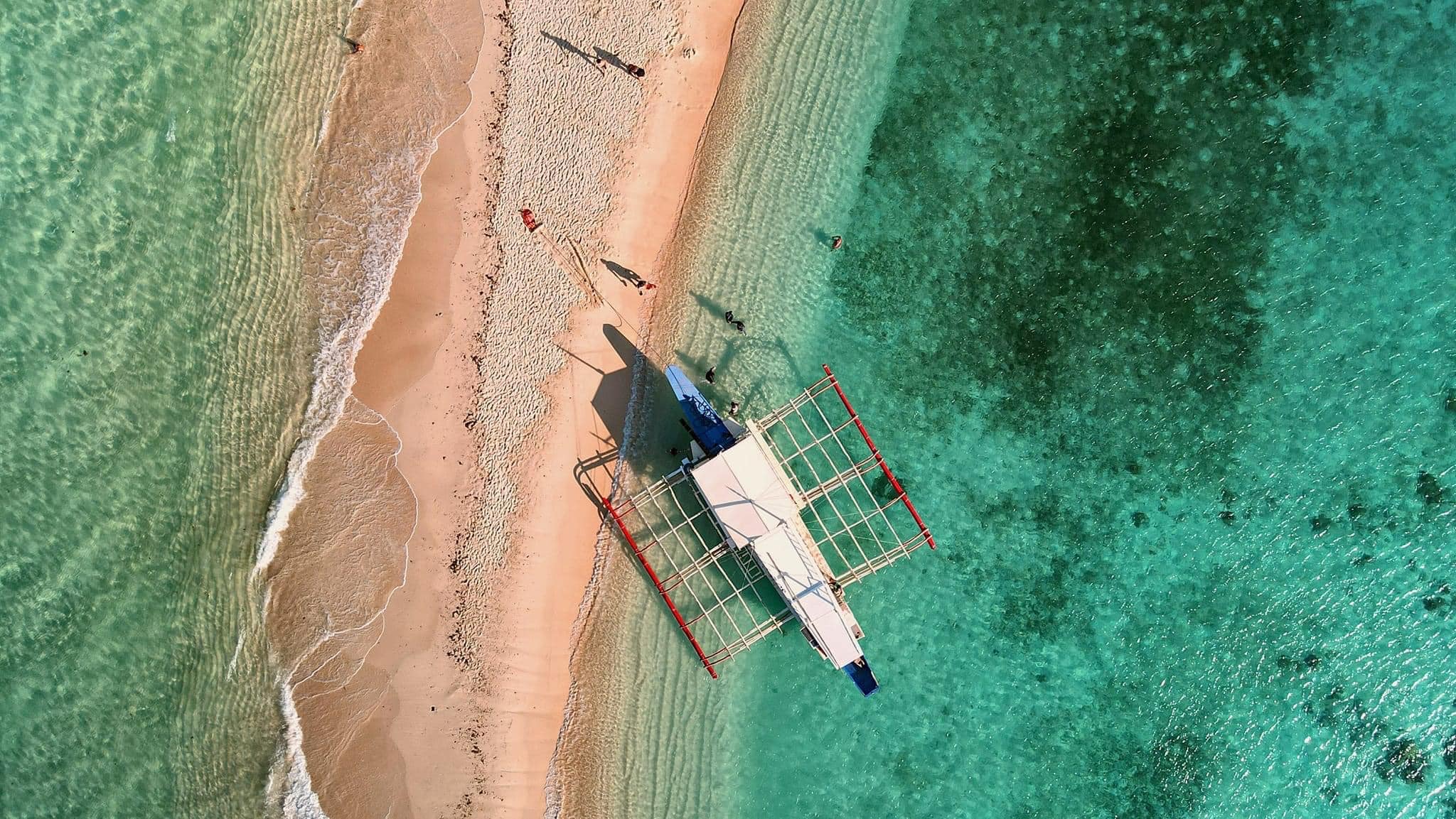 Aerial view of a traditional boat anchored on the narrow sandy shores of Seven Commando Beach in Palawan, surrounded by turquoise waters. People stroll along the shorelines of El Nido, where the sea is crystal clear with visible underwater features.