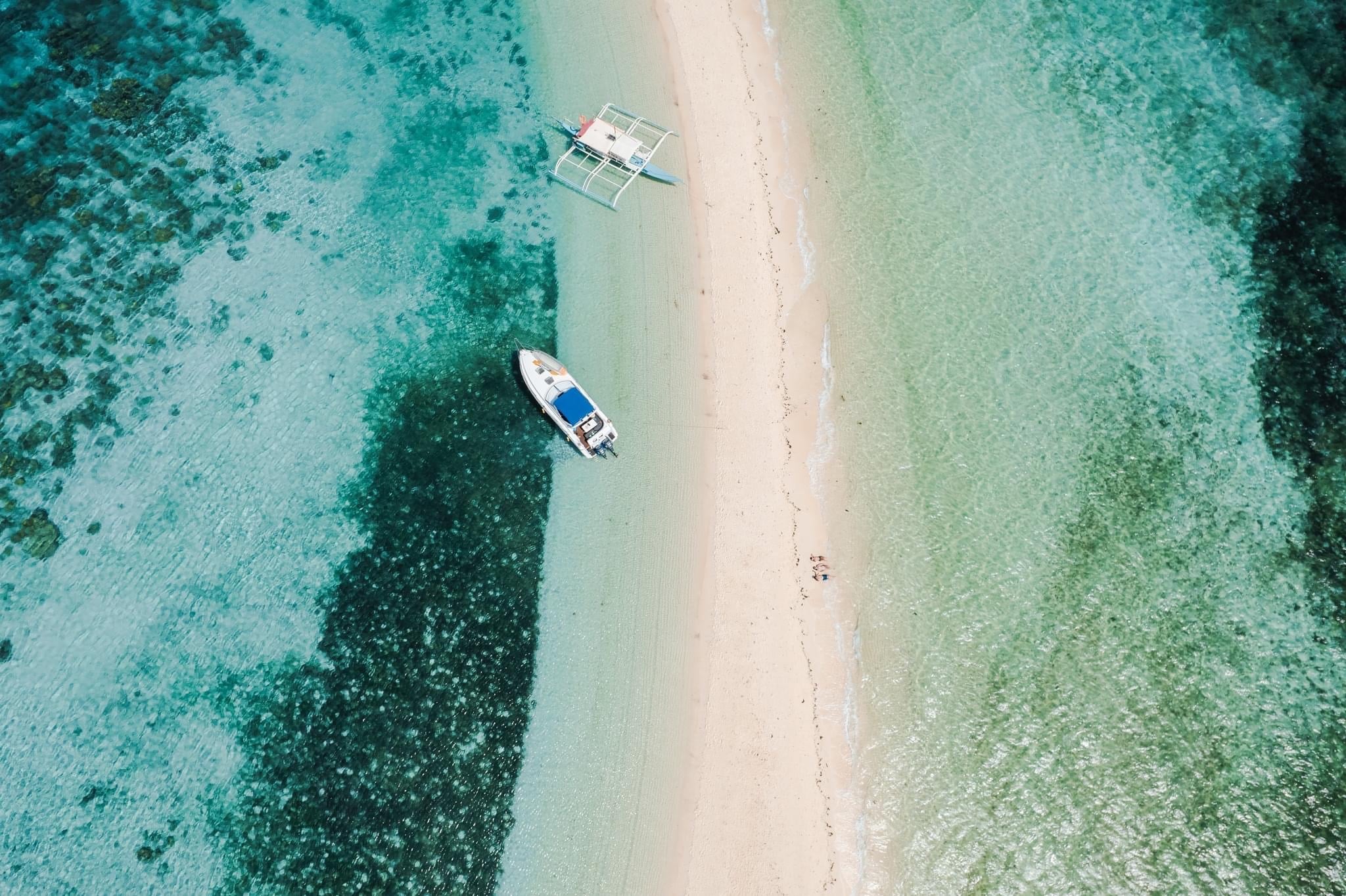 Aerial view of a narrow sandbar surrounded by the turquoise and deep blue waters of Coron, Palawan, with two boats anchored. Dark patches of coral are visible beneath the water near Ditaytayan Island as the sandbar gradually narrows stretching into the distance.