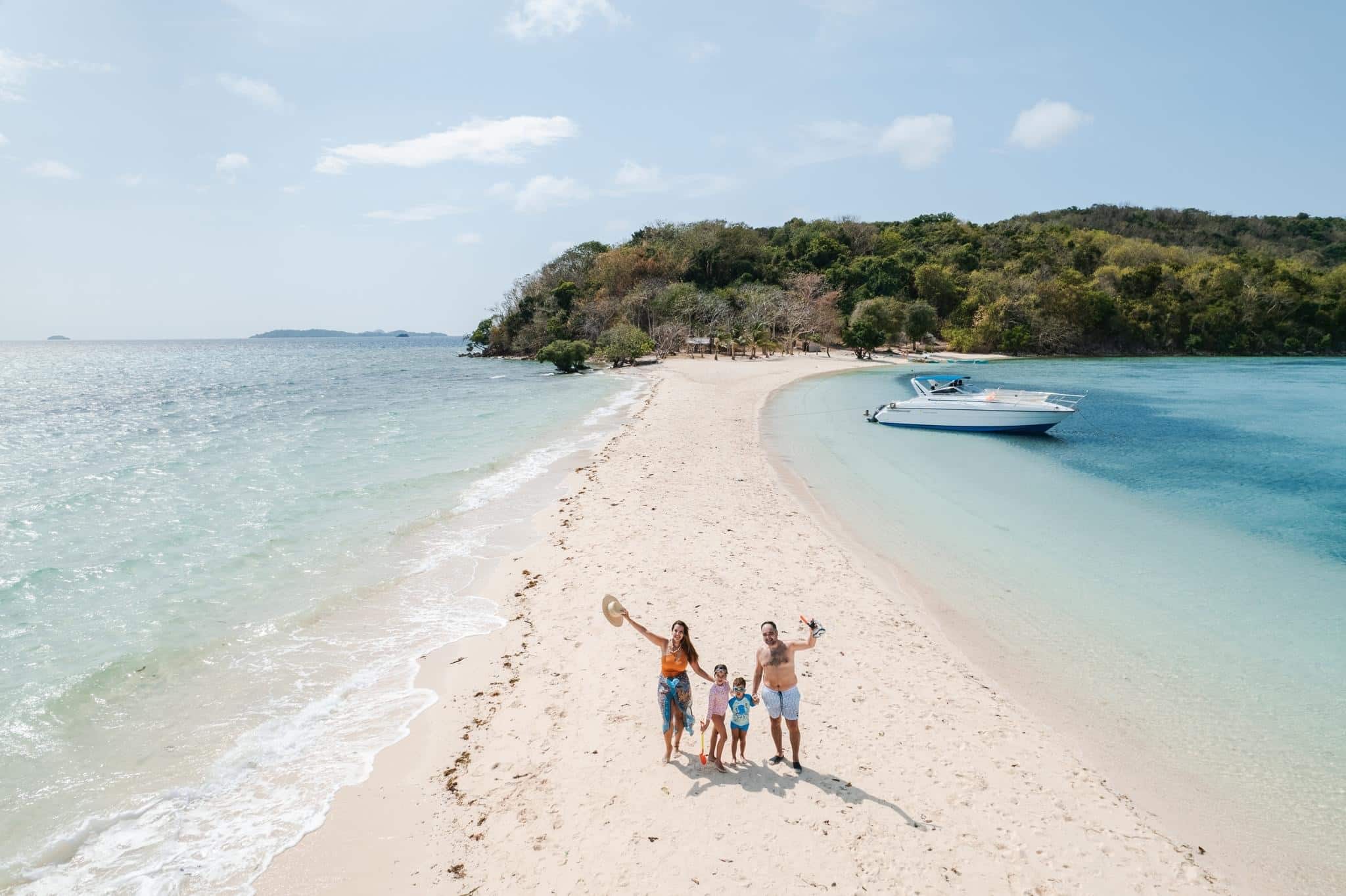 A family of four stands on a narrow sandy beach with clear turquoise water on both sides, waving cheerfully at the camera. With a boat docked nearby and Ditaytayan Island in Coron, Palawan providing a picturesque backdrop under the sunny sky, they capture a perfect seaside moment.