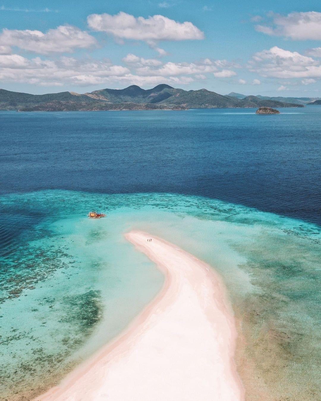Aerial view of a stunning, isolated sandy beach on Ditaytayan Island, Palawan, surrounded by vibrant turquoise waters and coral reefs. Distant green hills of Coron and islands under a blue sky with scattered clouds complete the breathtaking scene.