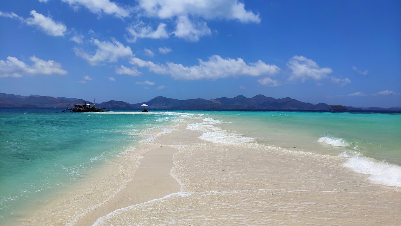 A scenic view of a narrow sandy beach on Ditaytayan Island in Coron, Palawan, surrounded by turquoise water under a clear blue sky. In the distance, small boats glide past lush green hills. Sparse, white clouds enhance the serene atmosphere of this tropical seaside paradise.