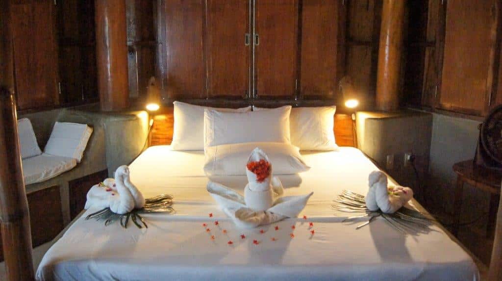 A cozy resort bed with white linens features two towel swans facing each other, while a third towel stands adorned with red flowers. This charming accommodation choice is framed by warm wooden walls and small side lights, perfect after a day of island hopping.