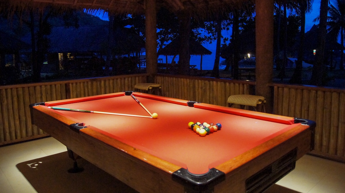 A red-felt pool table with billiard balls arranged for a game. Two cue sticks rest on the table under the dimly lit, thatched roof. This peaceful setting is perfect after a day of island hopping, offering a twilight view with trees silhouetted against the sky.