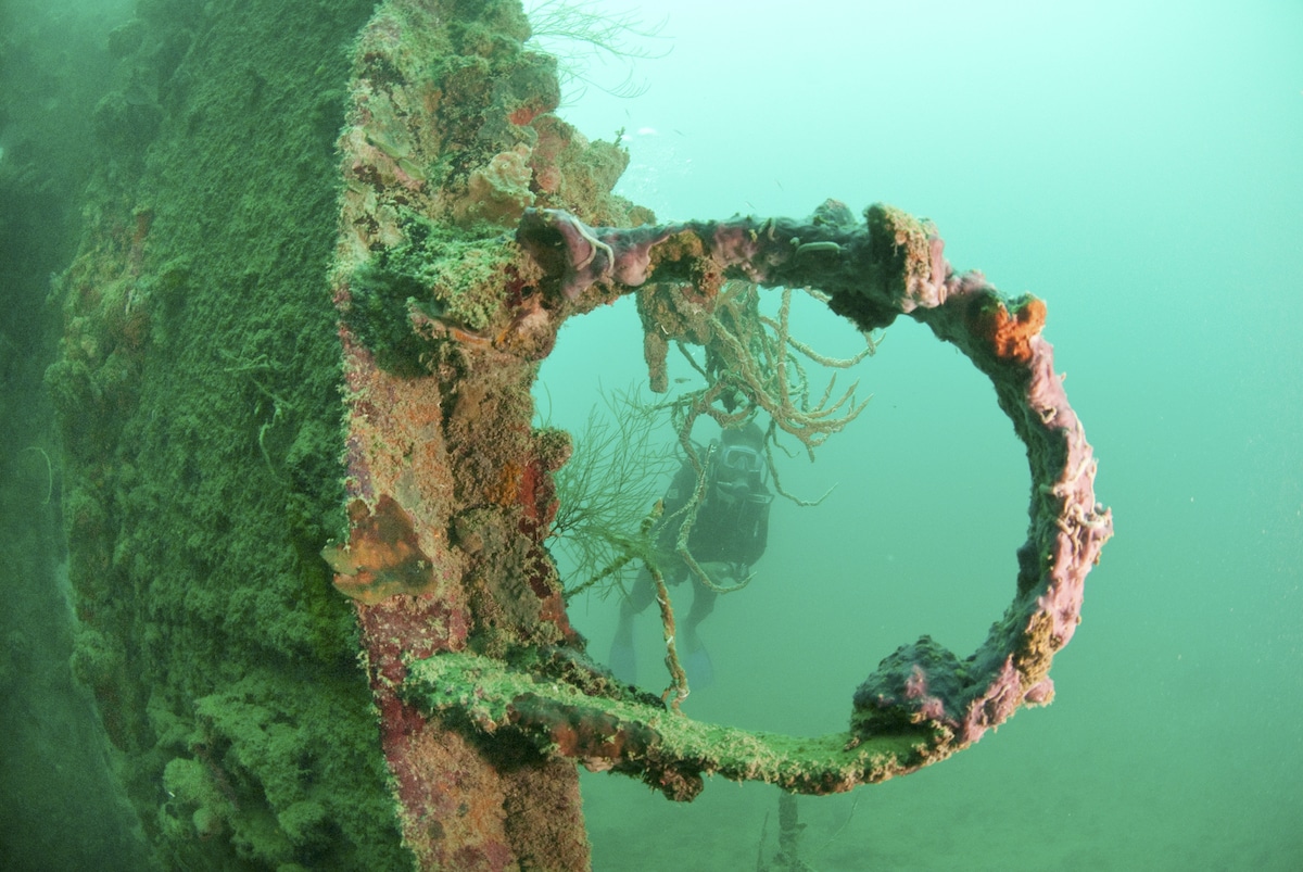 A diver explores an underwater shipwreck, a popular attraction for resort guests on boat tours. Marine growth and coral envelop the rusted circular structure in the foreground, highlighting the ship's skeletal remains against a greenish-blue ocean backdrop.