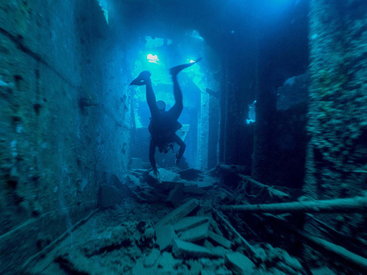 A scuba diver explores the interior of an underwater shipwreck, a hidden gem often discovered during the boat tour, surrounded by scattered debris and submerged structures. The scene is bathed in a blue hue from the ocean water spear fishing.