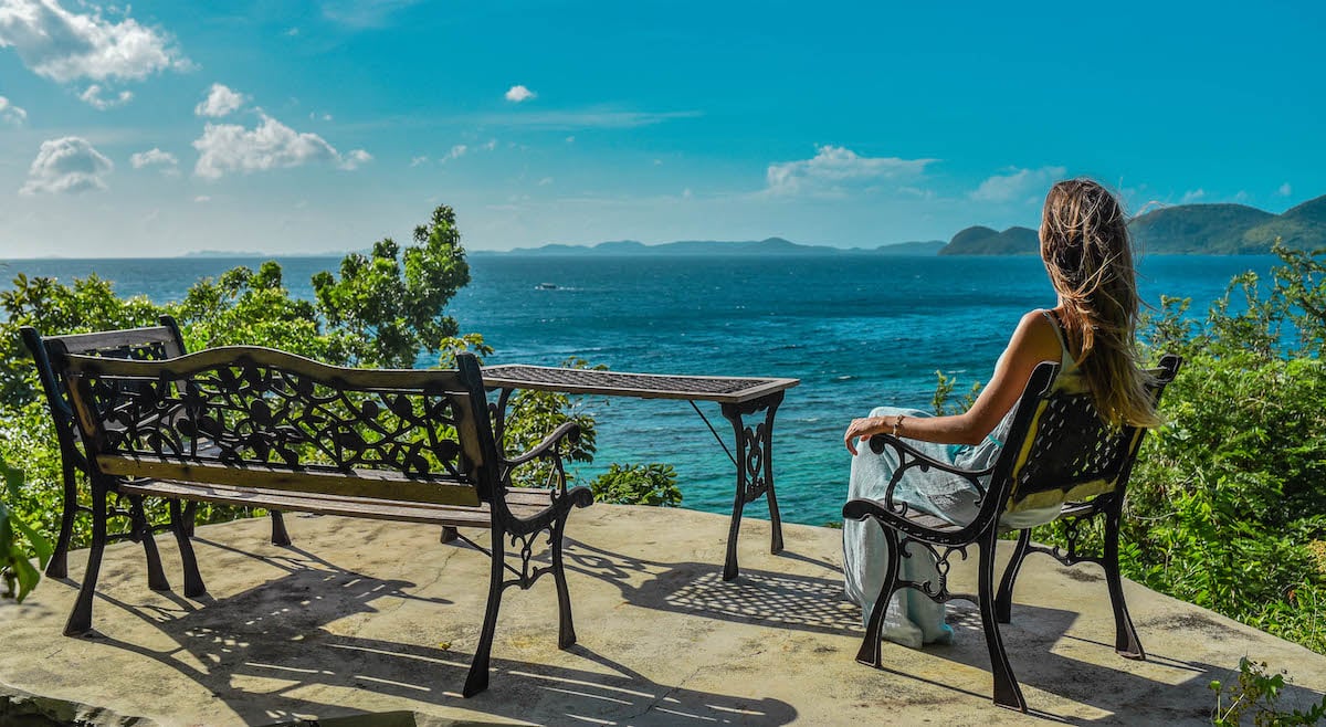 A person with long hair sits on a wrought iron bench, overlooking a vast ocean under a clear blue sky. The scene includes lush greenery and distant islands perfect for island hopping, evoking a sense of tranquility and scenic beauty.