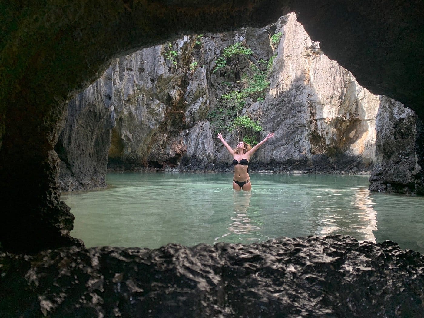 A person in a bikini stands joyfully with arms raised in El Nido's Secret Lagoon, nestled within a secluded, rocky cove framed by a cave opening, surrounded by lush greenery and calm water.