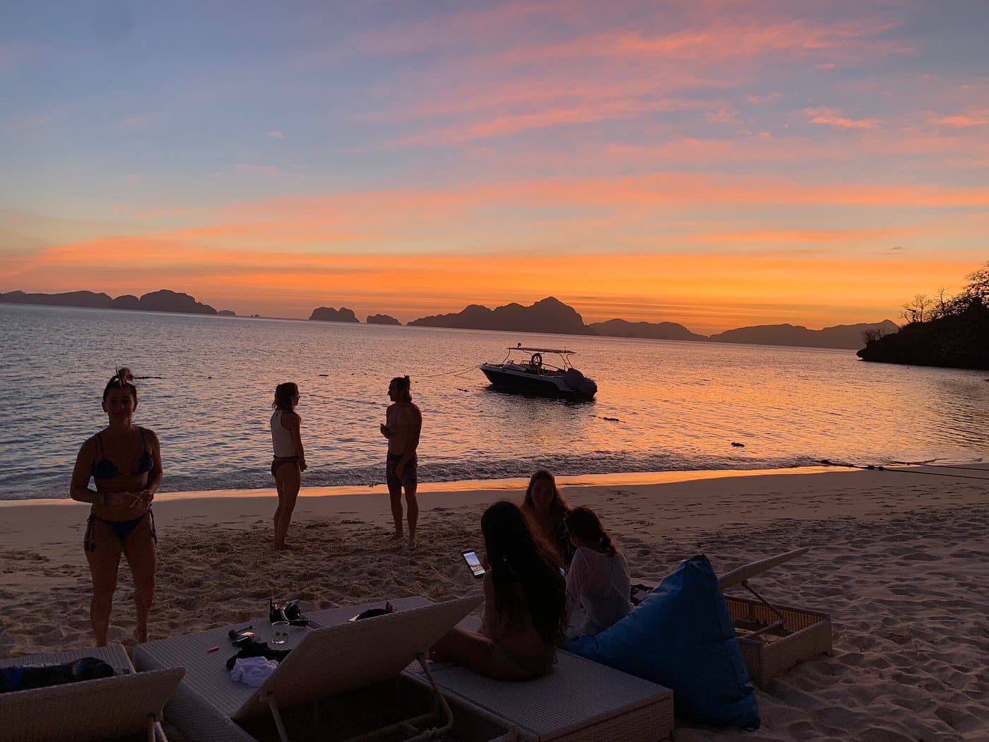 A group of people relax on Seven Commando Beach in El Nido at sunset, with chairs and towels arranged near the shore. A boat is anchored on the calm sea, and distant islands in Palawan are silhouetted against the vibrant orange and pink sky.