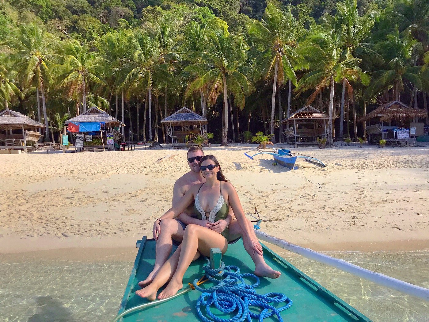 A couple sits at the front of a small boat on clear water at Seven Commando Beach, with a sandy shore and palm trees framing the scene in El Nido, Palawan. Several rustic beach huts peek through the trees, adding to the idyllic charm.