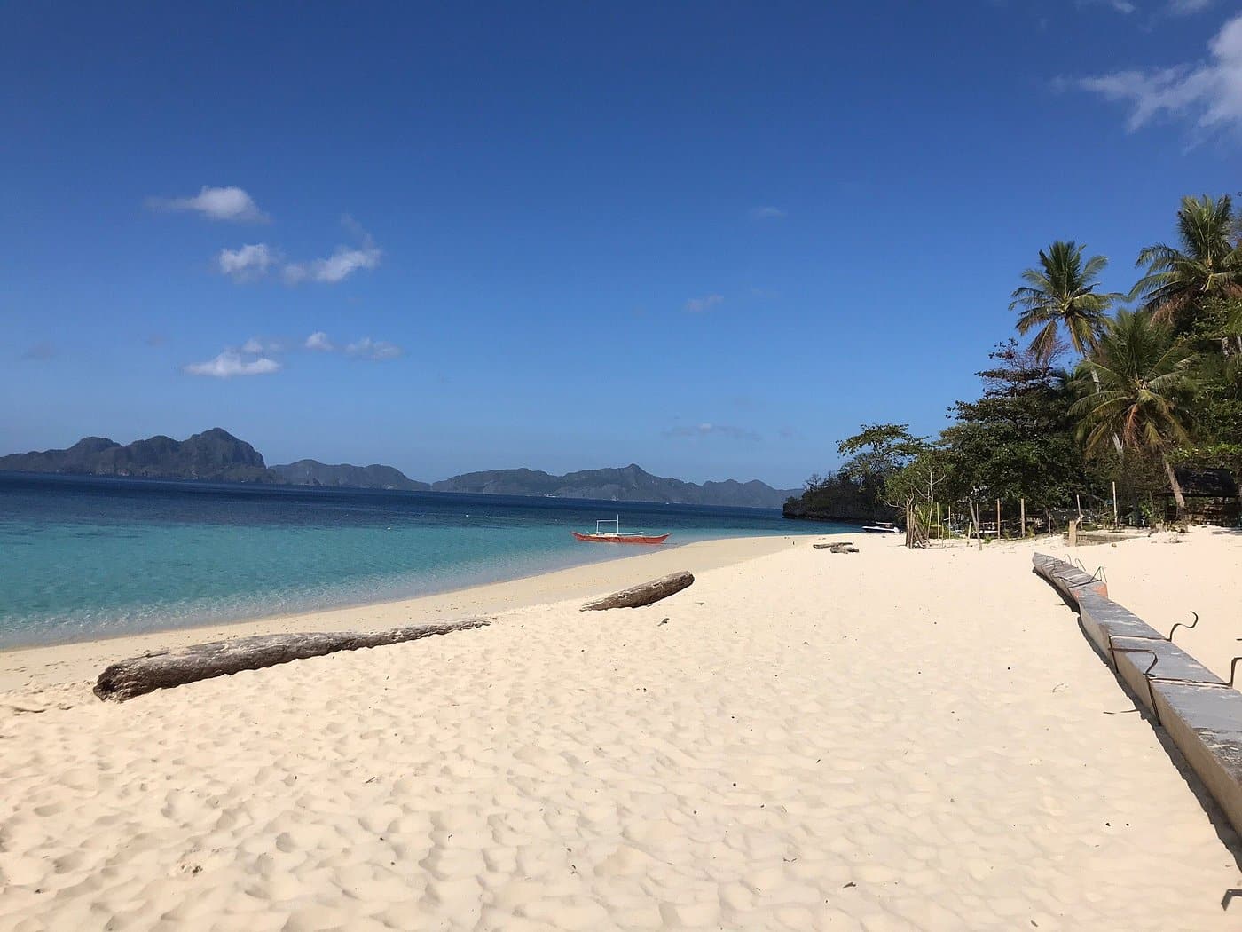 A serene tropical beach with white sand, clear blue water, and a small boat in the distance. Lush green trees line the shore of Seven Commando Beach in El Nido, Palawan, with distant islands visible under a clear sky. Long logs are scattered on the sand.