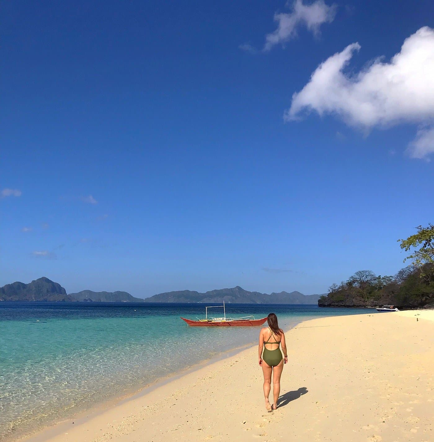 A person in a swimsuit strolls along the sandy Seven Commando Beach in Palawan, heading toward a boat on clear turquoise waters, surrounded by distant islands under a blue sky dotted with a few clouds.