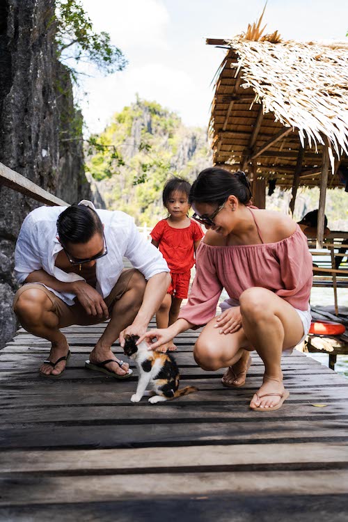 On a sunny day, a family of three, with a small child, pauses their tour to bend down and pet a calico cat on a wooden dock. They are near a thatched hut, surrounded by lush greenery and shimmering waters.