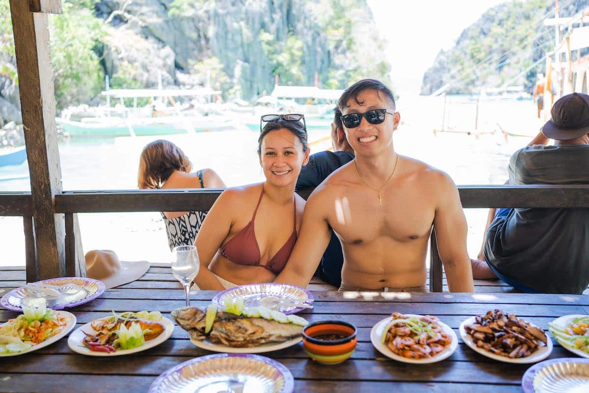 A couple in swimwear sits at a table laden with various dishes, including grilled seafood and vegetables, overlooking a scenic water view. As they smile and soak up the sun, it seems like the perfect setting for a marriage proposal against the backdrop of boats and rocky cliffs.