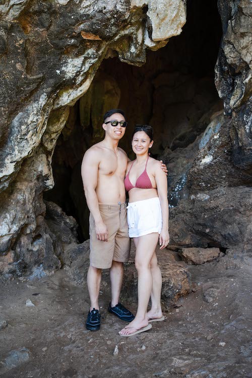 A smiling couple stands at the entrance of a rocky cave. The man is shirtless, wearing sunglasses and brown shorts, while the woman is in a red bikini top and white shorts. They appear to be savoring a sunny day outdoors, possibly on a tour or celebrating their recent marriage proposal.