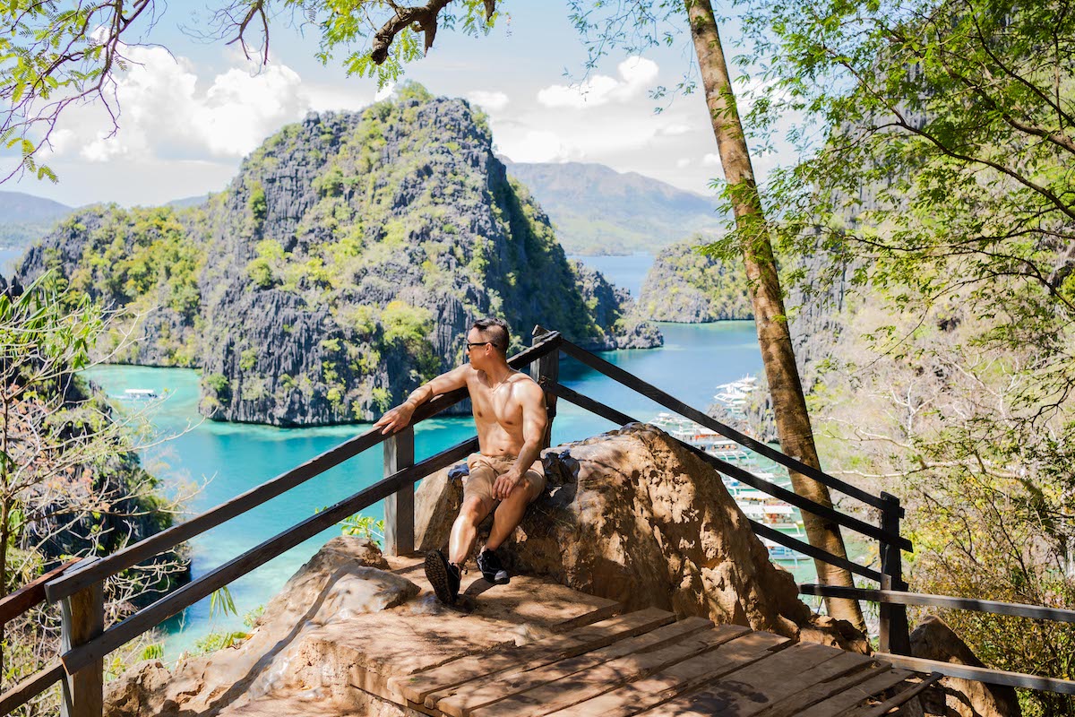 A person sits on a wooden platform overlooking a turquoise bay with steep, lush cliffs under a clear sky, perhaps contemplating the perfect spot for a marriage proposal. The scenic view is surrounded by tropical trees and plants, creating a vivid, natural landscape ideal for memorable tours.