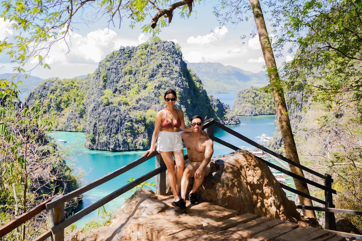 A couple poses on a wooden lookout, perhaps after a heartfelt proposal, with turquoise waters and limestone cliffs in the background. The clear, sunny sky and green foliage frame their moment beautifully.