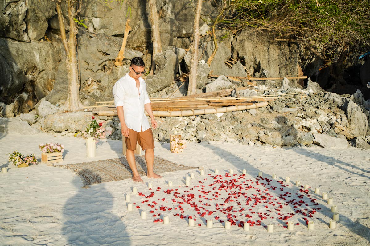 A person stands on a sandy beach, ready for a marriage proposal. They're surrounded by a heart-shaped arrangement of red petals and candles, with flowers and a wooden crate nearby. Rocky cliffs and trees create a stunning backdrop for this romantic moment.