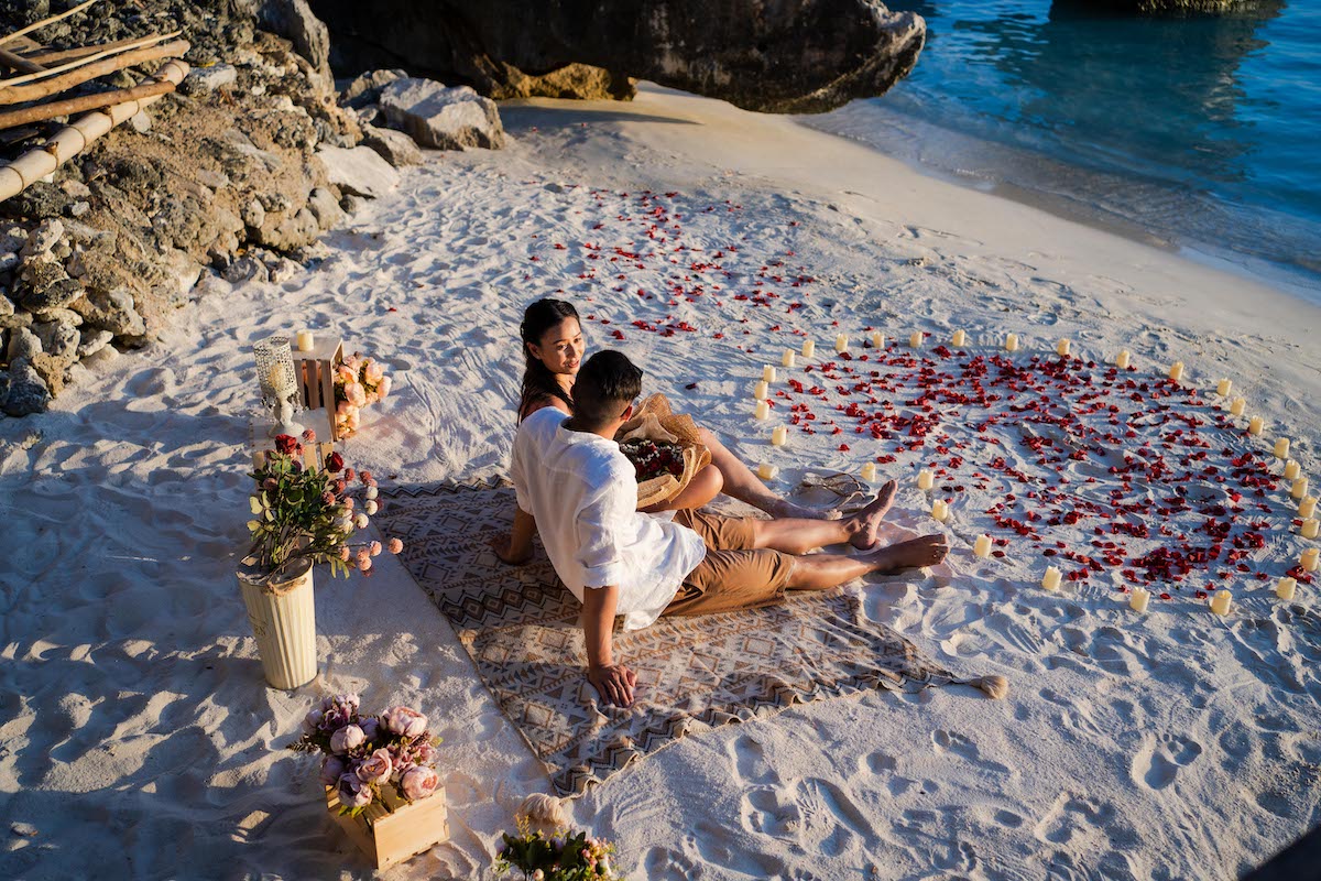 A couple sits on a patterned blanket at the beach, surrounded by rose petals arranged in a heart shape, decorative flowers, and candles—the perfect setting for a surprise proposal. The ocean and rocks provide a stunning backdrop under the clear sky.