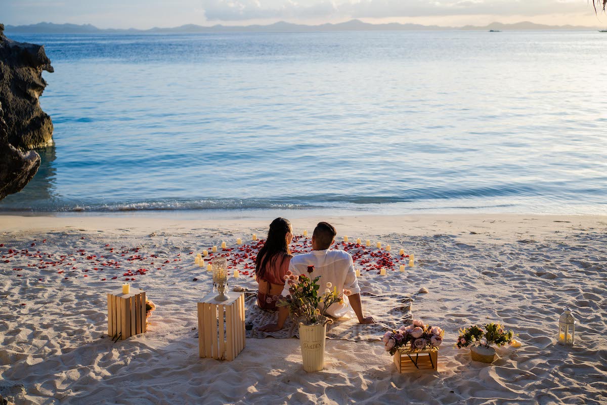 A couple sits on a sandy beach, surrounded by scattered petals and floral arrangements, gazing out at the tranquil ocean under a soft sunset sky. Wooden crates and candles hint at an unforgettable proposal in this romantic setting.