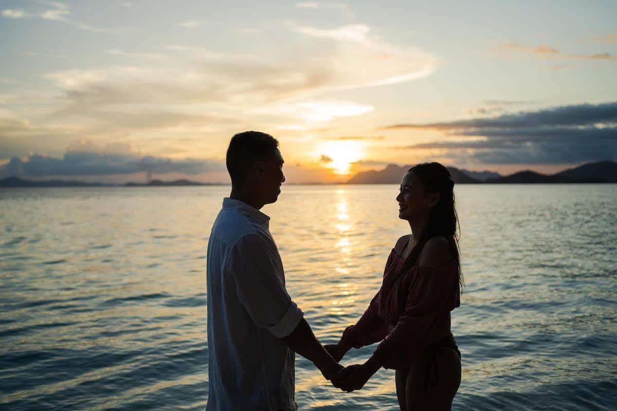 A couple stands holding hands at the beach during sunset, perfect for a romantic proposal. Their silhouettes are set against a backdrop of calm sea and a colorful sky, with hues of orange and pink reflecting on the water.