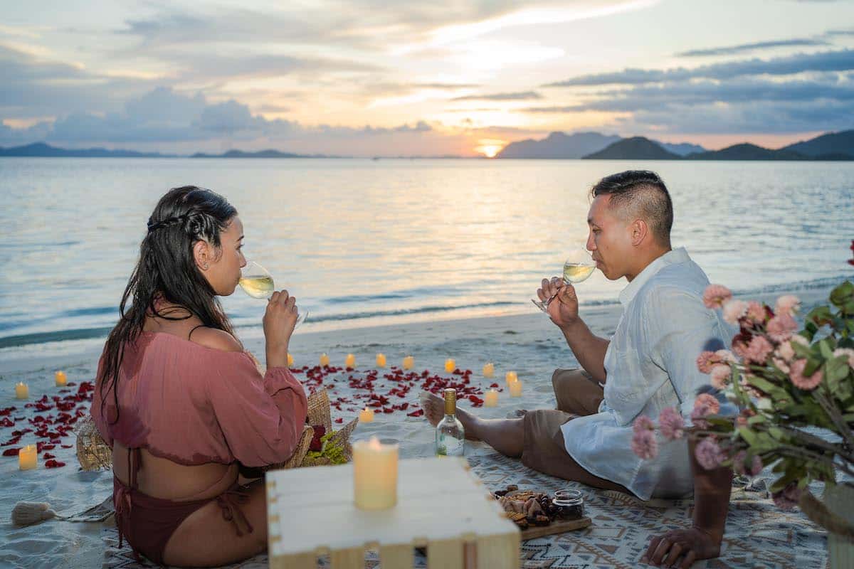 A couple enjoys a romantic beach picnic during sunset, with drinks in hand. They sit on a blanket surrounded by rose petals and candles, perfect for a surprise proposal overlooking the calm sea. In the foreground, there's a small table with snacks and a flower arrangement.