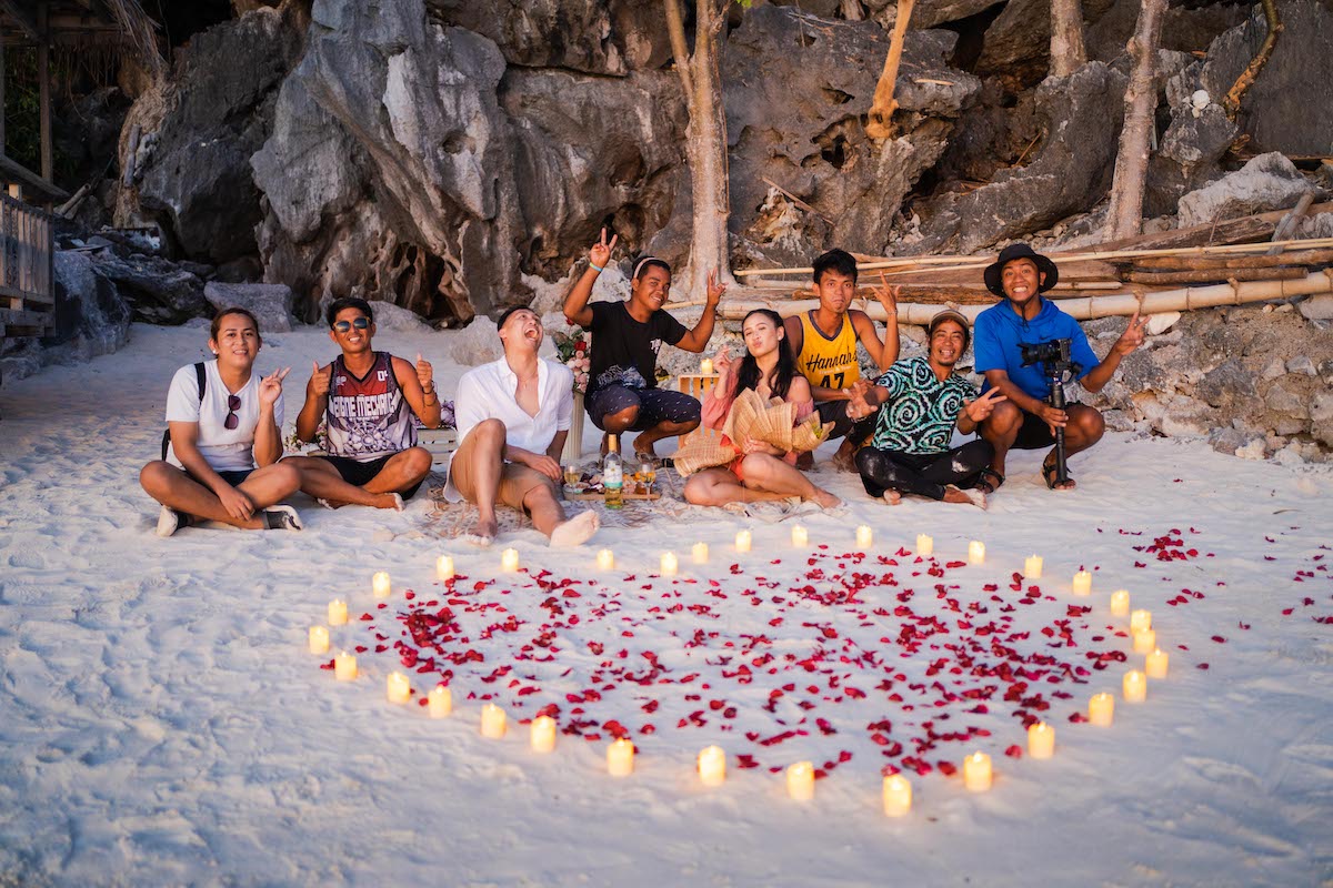 Eight friends sit joyfully on a beach, encircled by a heart of rose petals and candles, capturing the moment with rocky cliffs and trees framing their joy. It's a perfect setting for an unexpected marriage proposal amid nature's backdrop.