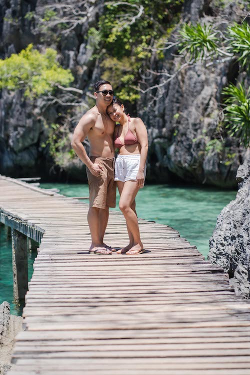 A couple stands on a wooden bridge over turquoise water, ready for a proposal. The man is shirtless, wearing sunglasses and shorts, while the woman wears a bikini top and white shorts. Lush greenery and rocky cliffs create the perfect backdrop for this memorable moment.
