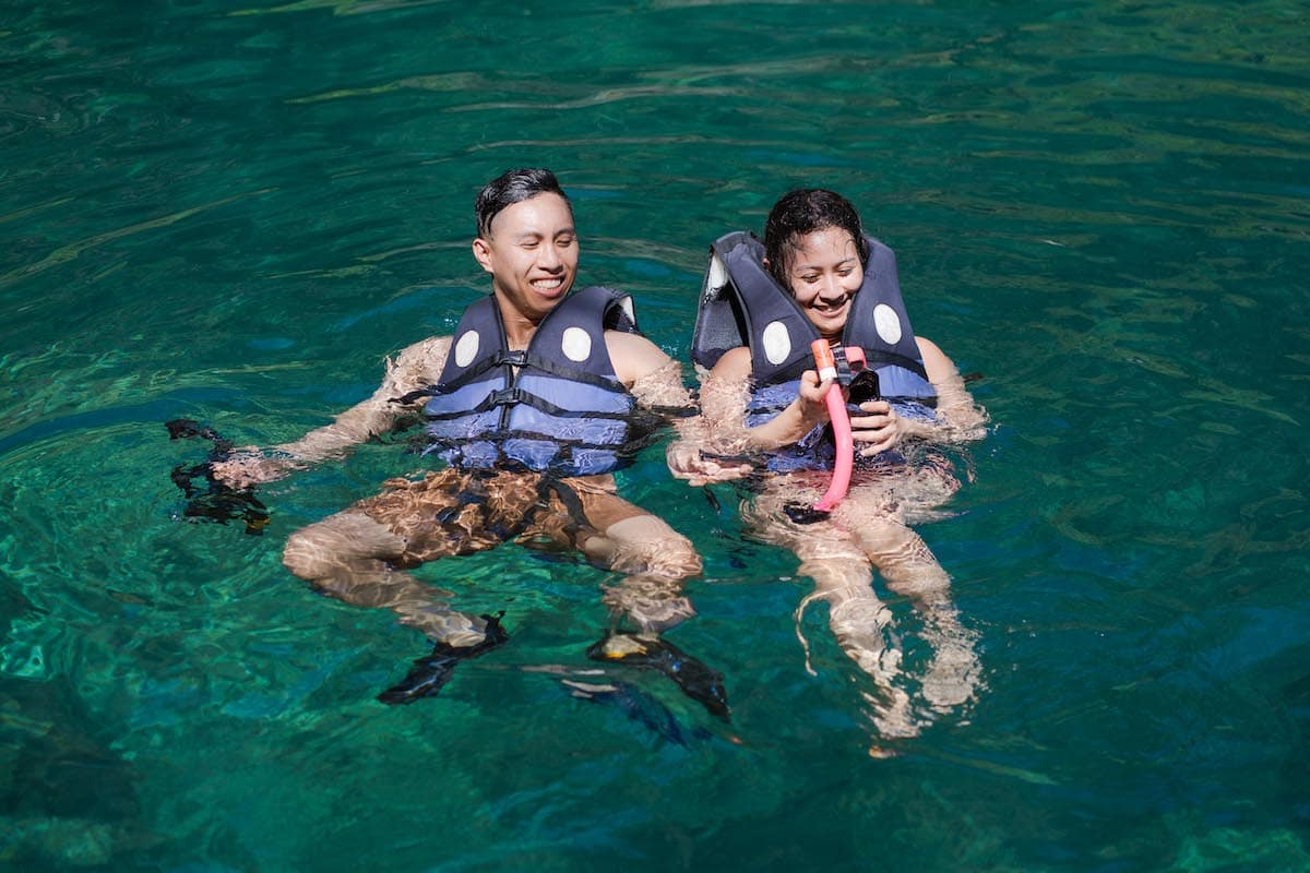 Two people wearing life vests are floating in clear, green water, smiling and enjoying the sunny day. One holds a small camera with a red strap, perhaps capturing the perfect moment of their tour.