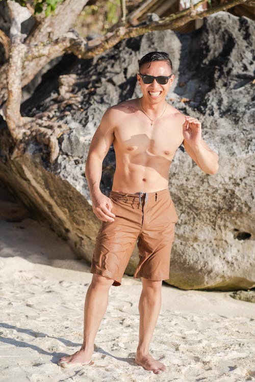 A shirtless man wearing sunglasses and brown swim trunks grins while posing on a sandy beach, perhaps contemplating a proposal. Large rocks and lush green foliage form a stunning backdrop perfect for a memorable tour.