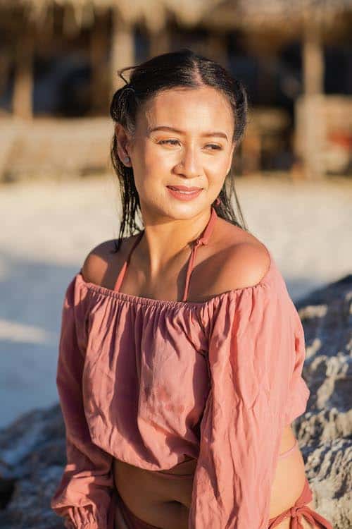 A person with long dark hair smiles while sitting on a rock at the beach, wearing an off-the-shoulder pink top. The sunlight casts a warm glow on their face, creating the perfect backdrop for a surprise marriage proposal. The background features blurred sand and a wooden structure.