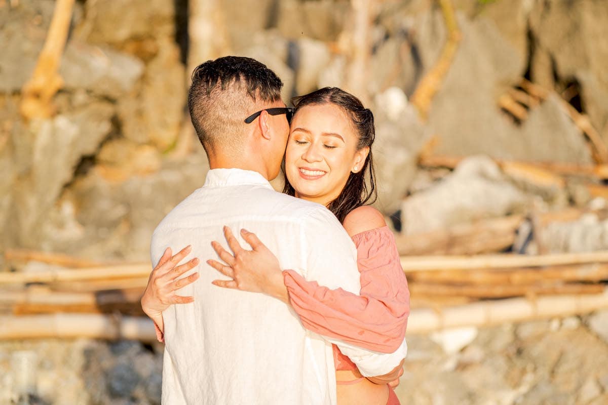 Amidst a sunlit rocky backdrop, a woman in a pink off-shoulder dress embraces a man in a white shirt and sunglasses. With her eyes closed in bliss, she smiles warmly. Could this tender moment mark the proposal that begins their journey toward marriage?