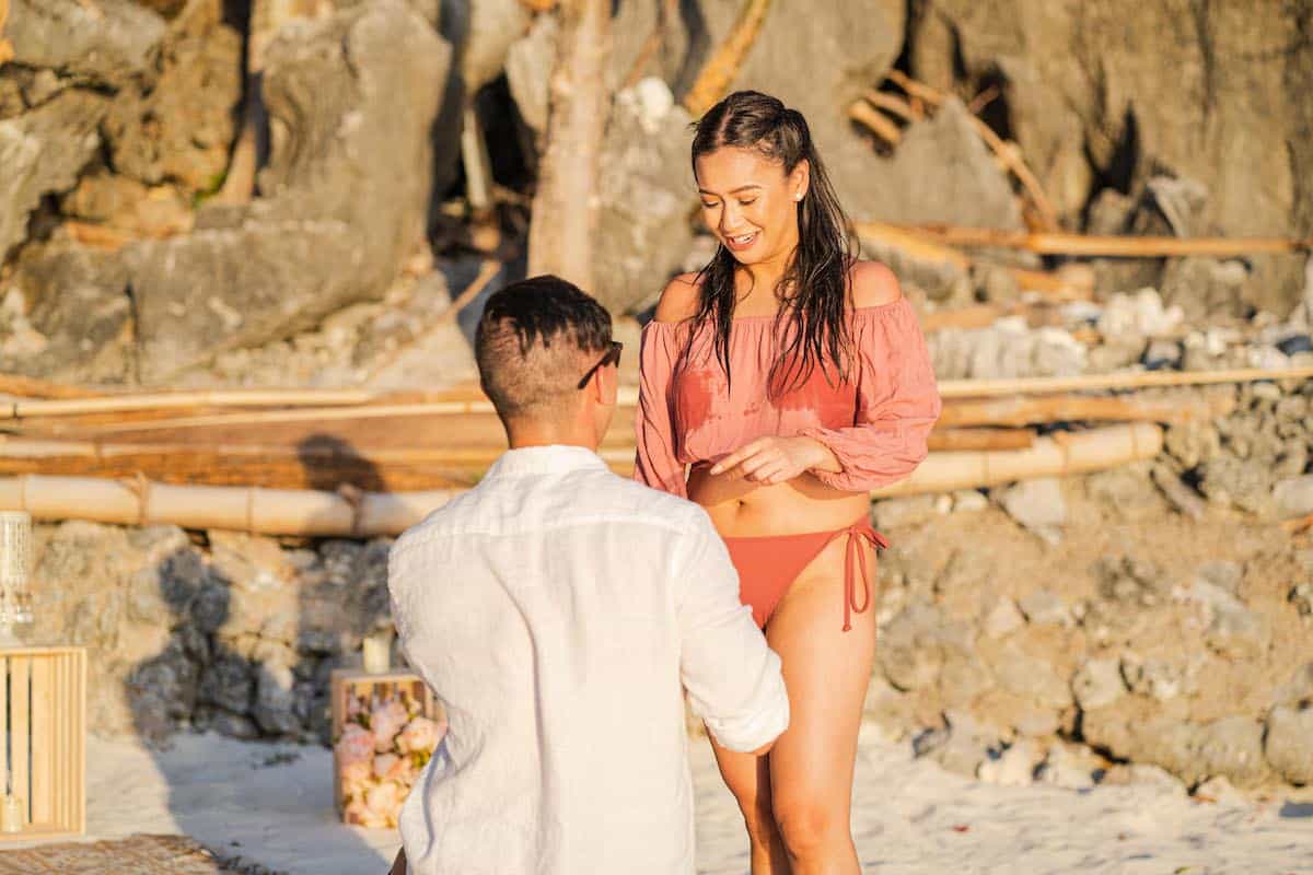 A man kneels on one knee on a sandy beach, holding a ring box in a heartfelt proposal. A woman in a pink blouse and bikini bottoms smiles at him amid the rocky terrain and beach decor. The scene beautifully captures their journey toward marriage.