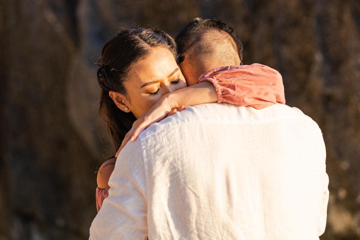 A couple embraces tenderly, with the woman's eyes closed, resting her head on the man's shoulder—a serene moment possibly marking a touching marriage proposal. They are both wearing light-colored clothing, and the background is softly blurred, enhancing their intimacy.