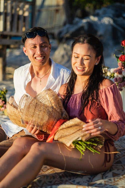 A smiling couple enjoys a sunny beach setting, possibly after a proposal. The woman holds a bouquet wrapped in rustic paper. Seated on a patterned blanket with flowers in the background, it suggests a romantic or celebratory occasion.